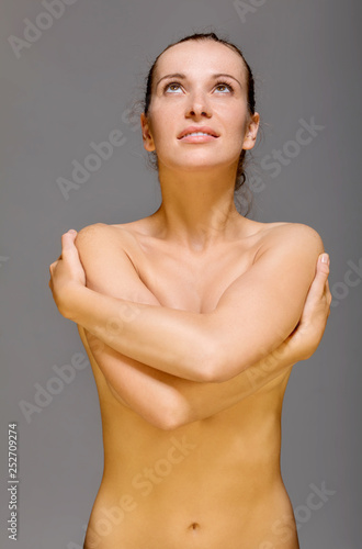 Naked woman covering her breast and looking up, on grey background.