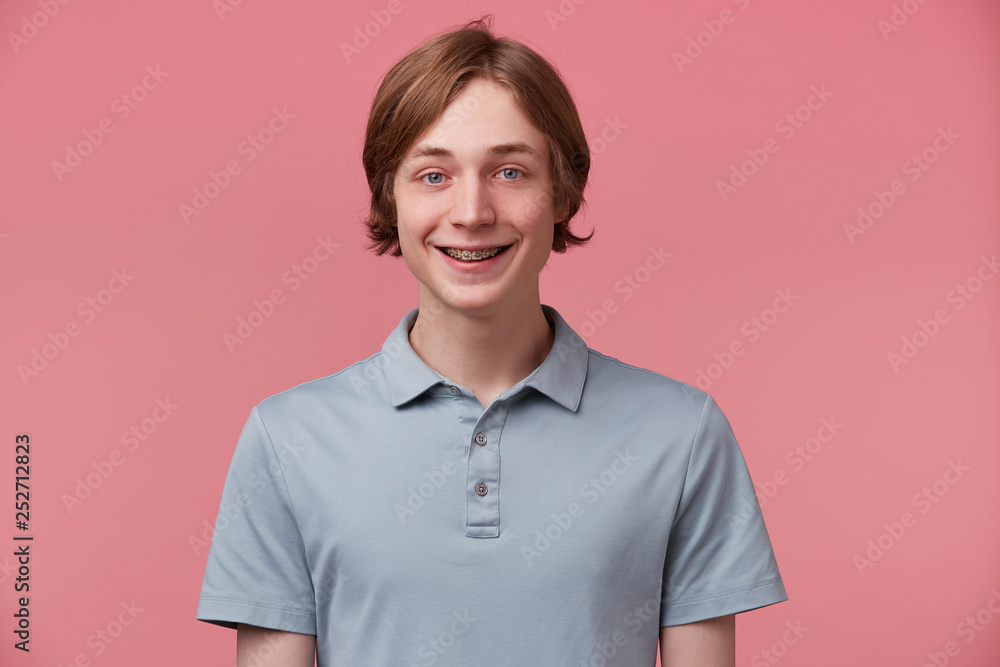 Close up of blue-eyed neatly combed young guy with braces on teeth wears polo t-shirt slightly smiles and feels happy isolated over pink background Close up of blue-eyed neatly combed young guy with braces on teeth wears polo t-shirt slightly smiles and feels happy isolated over pink background
