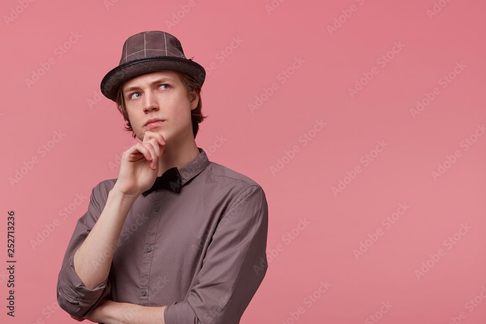 Young elegantly dressed guy stands holding his hand on a chin, looking right upper corner thoughtfully, serious, thinking about art, ponders about some literature, over pink background Young elegantly dressed guy stands holding his hand on a chin, looking right upper corner thoughtfully, serious, thinking about art, ponders about some literature, over pink background
