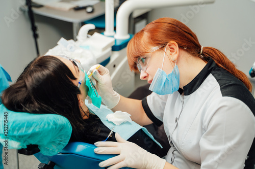 Red-haired dentist wearing mask, gloves and uniform examining a patient teeth in clinic. Stomatologist in glasses treats female teeth with medical equipment.