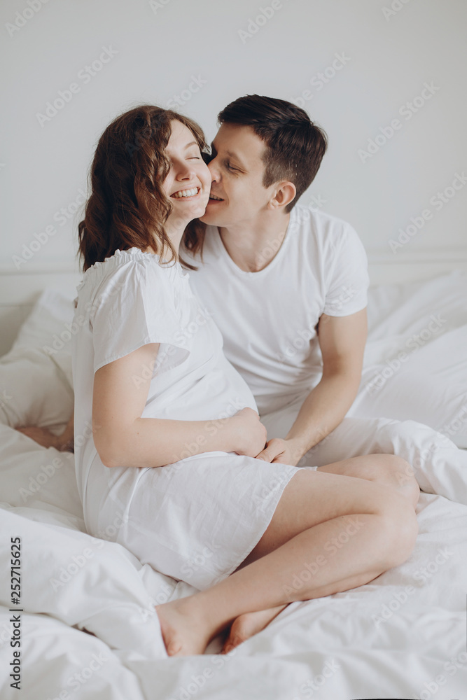 Happy pregnant couple relaxing on white bed and holding belly bump. Happy young husband kissing his smiling wife and hugging baby belly. Stylish pregnant family in white at home. True happiness Happy pregnant couple relaxing on white bed and holding belly bump. Happy young husband kissing his smiling wife and hugging baby belly. Stylish pregnant family in white at home. True happiness