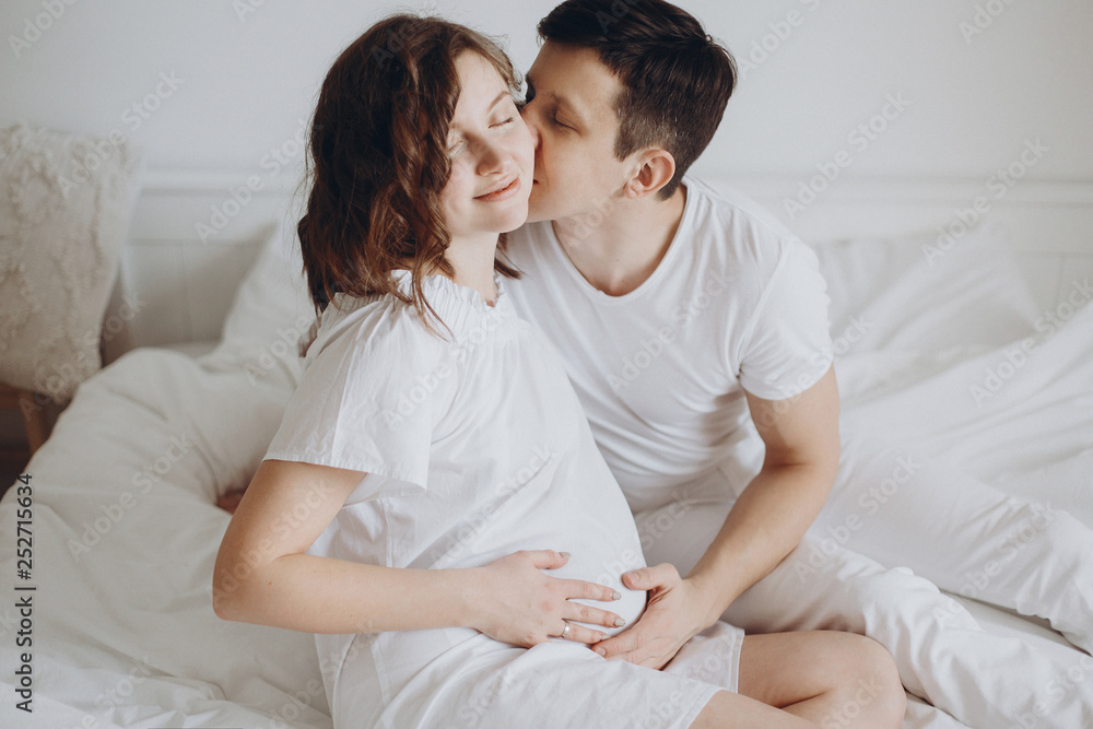 Happy young husband kissing his smiling pregnant wife and hugging belly bump on white bed. Stylish pregnant couple in white relaxing and holding belly at home. Fertility concept. True happiness Happy young husband kissing his smiling pregnant wife and hugging belly bump on white bed. Stylish pregnant couple in white relaxing and holding belly at home. Fertility concept. True happiness