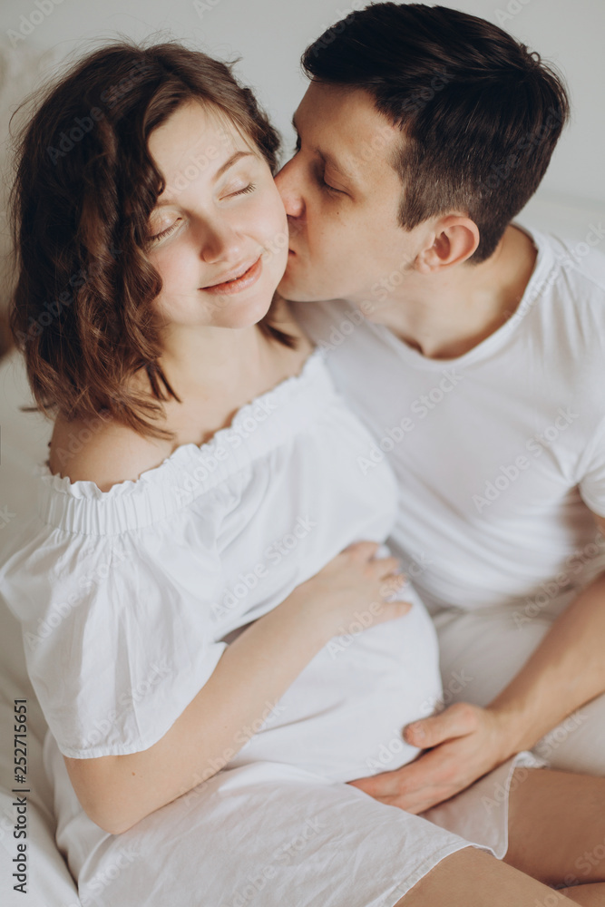 Happy pregnant couple relaxing on white bed and holding belly bump. Happy young husband kissing his smiling wife and hugging baby belly. Stylish pregnant family in white at home. True happiness Happy pregnant couple relaxing on white bed and holding belly bump. Happy young husband kissing his smiling wife and hugging baby belly. Stylish pregnant family in white at home. True happiness