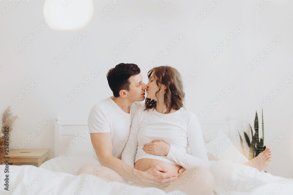 Happy young husband kissing his smiling pregnant wife and hugging belly bump on white bed. Stylish pregnant couple in white relaxing and holding belly at home. Fertility concept. True happiness Happy young husband kissing his smiling pregnant wife and hugging belly bump on white bed. Stylish pregnant couple in white relaxing and holding belly at home. Fertility concept. True happiness