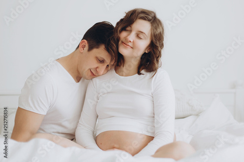 Happy young pregnant couple holding belly bump on white bed. Stylish pregnant family, mom and dad in white, relaxing at home and hugging belly. Fertility concept. Moment of true happiness