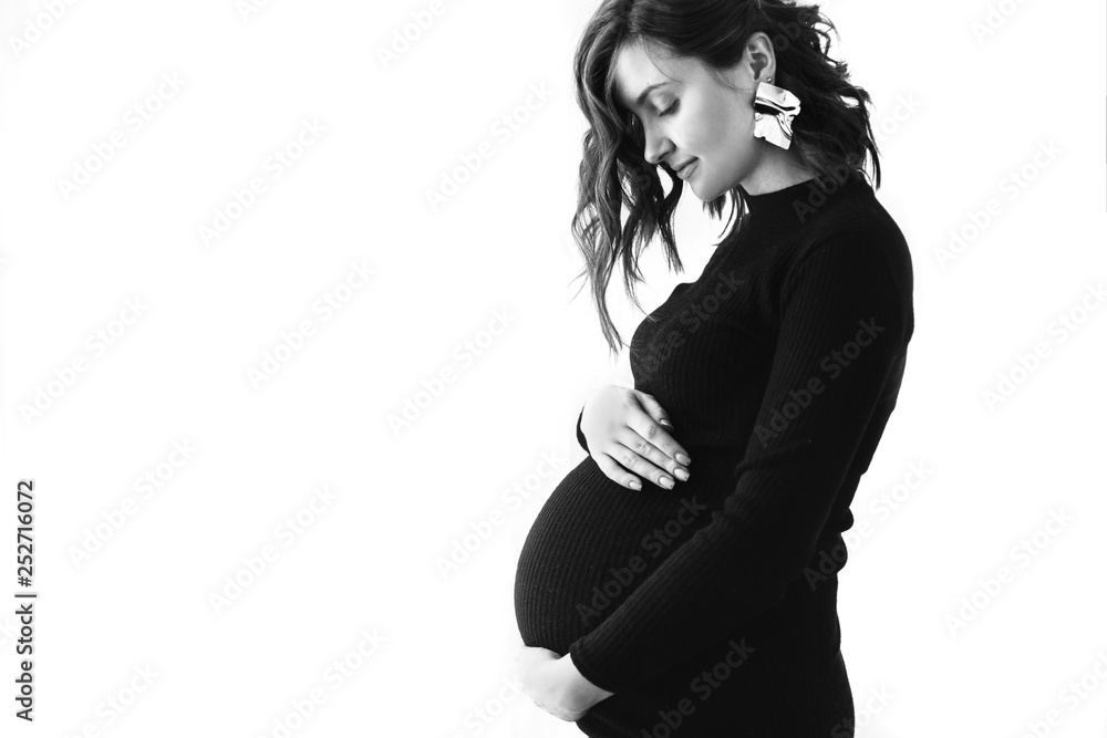 Fashionable pregnant mom, hugging belly. Stylish Motherhood concept. Space for text. Happy young pregnant woman in stylish black dress and modern earring, holding belly bump and posing in light Fashionable pregnant mom, hugging belly. Stylish Motherhood concept. Space for text. Happy young pregnant woman in stylish black dress and modern earring, holding belly bump and posing in light