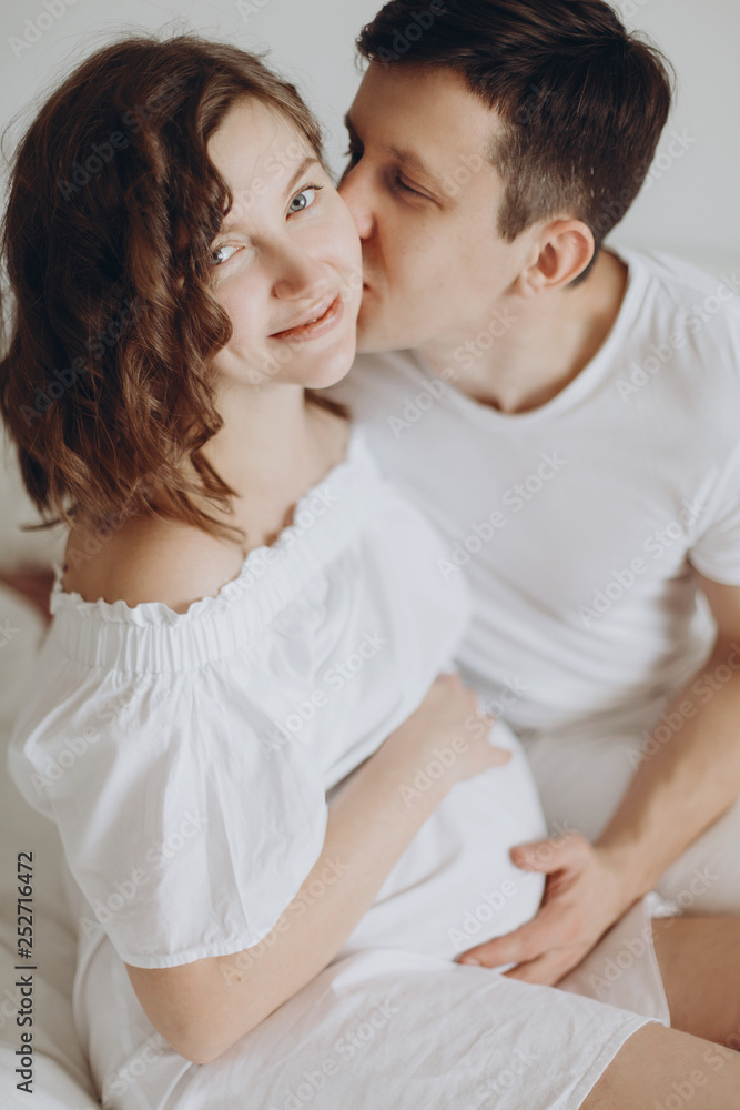 Happy young husband kissing his smiling pregnant wife and hugging belly bump on white bed. Stylish pregnant couple in white relaxing and holding belly at home. Fertility concept. True happiness Happy young husband kissing his smiling pregnant wife and hugging belly bump on white bed. Stylish pregnant couple in white relaxing and holding belly at home. Fertility concept. True happiness