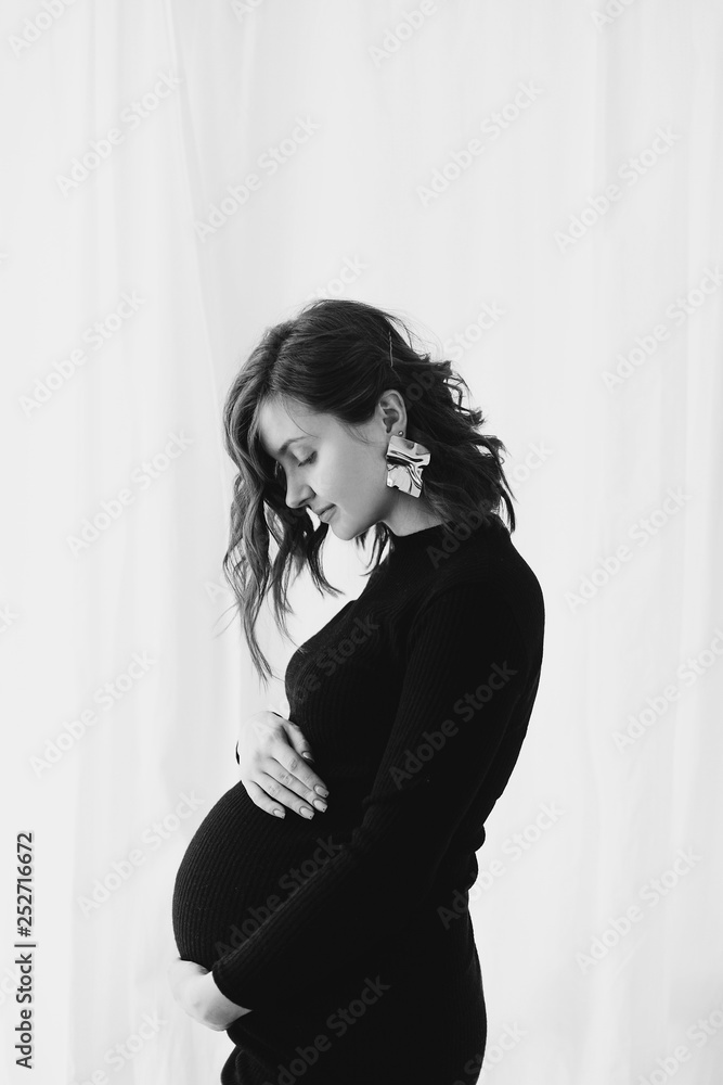 Fototapeta premium Happy young pregnant woman in stylish black dress holding belly bump and posing in light. Stylish fashionable mom, hugging belly, waiting for baby. Motherhood and fertility concept