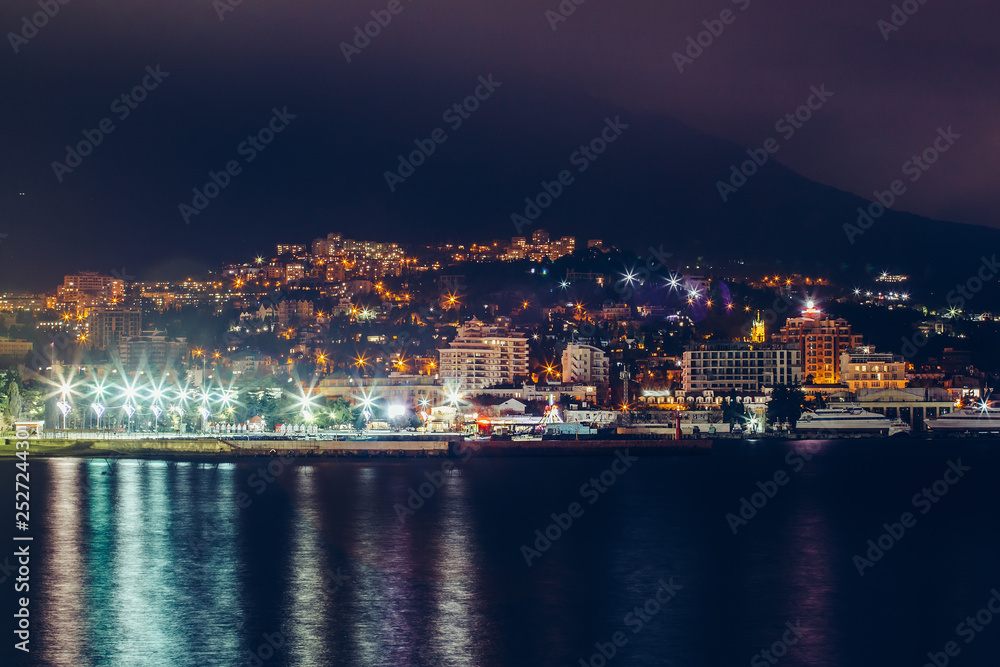 Yalta embankment at night, city buildings lights reflected in black water, beautiful resort in Crimea with mountains and sea