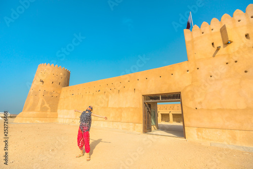 Travel in North of Qatar at Al Zubara Fort. Happy woman at entrance of old castle, a historic military fortress in Middle East, Arabian Peninsula. Blonde tourist in desert landscape of Persian Gulf.