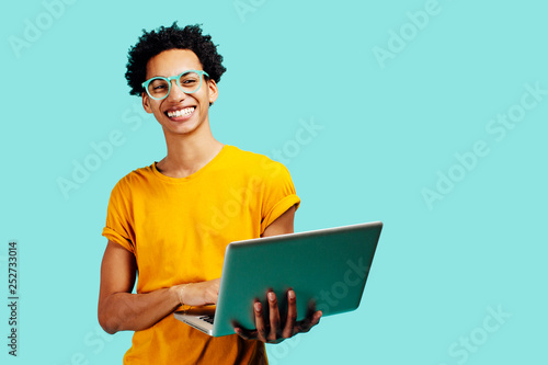 Portrait of a smiling young man with glasses holding his laptop, isolated on blue background