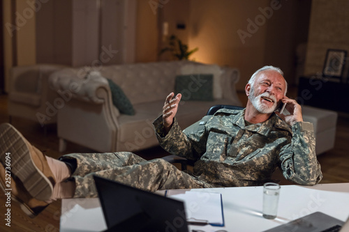 Cheerful military officer with feet up on table talking on cell phone.