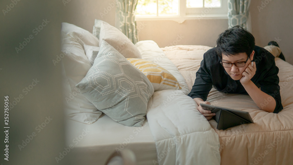 Young Asian business man using digital tablet and pen while lying on bed in cozy bedroom. Home living lifestyle with modern electronic gadget concept Young Asian business man using digital tablet and pen while lying on bed in cozy bedroom. Home living lifestyle with modern electronic gadget concept