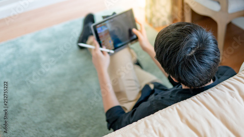 Young Asian business man using digital tablet and pen while sitting on carpet by the bed in cozy bedroom. Home living lifestyle with modern electronic gadget concept