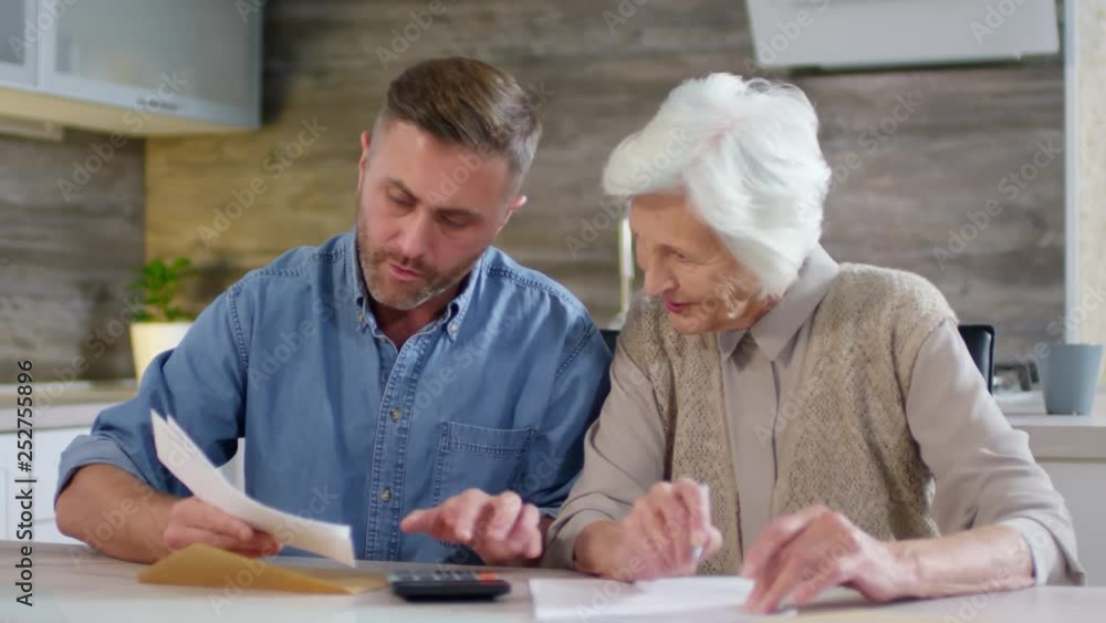 Medium shot of adult man and his aged mother sitting together at kitchen table and making calculations of family income Medium shot of adult man and his aged mother sitting together at kitchen table and making calculations of family income