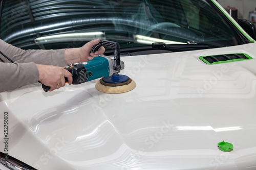 The master man of the detailing in work clothes and dirty hands polishes the bodywork of the bonnet of the car in white with a polishing machine in the workshop for body repair cars