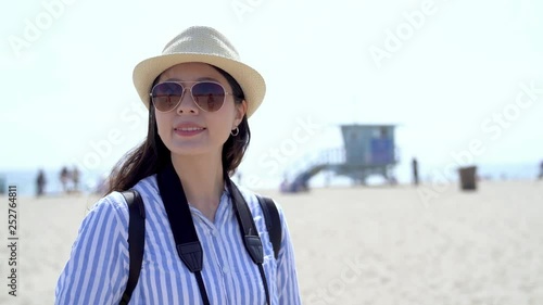 Young asian woman with hat and sunglasses walking on the beach in 
Santa Monica Pier relax under sunshine. beautiful lady traveler travel in usa self guide trip. happy girl carry camera sightseeing