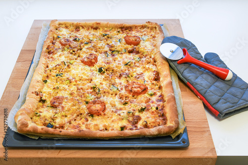 Freshly backed delicious crunchy homemade pizza with vegetables, tomatoe, corn, pumpkin on a rectangular metal tray and wooden table next to baking grey glove and red spinning knife
