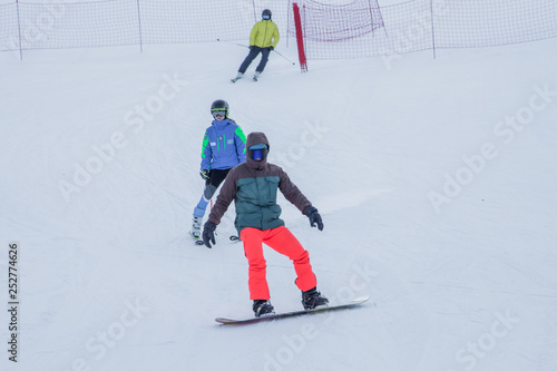 A snowboarder and two skiers running downhill on the white slope in the ski resort in Kazakhstan