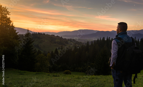 A bearded man (tourist) in a sleeveless jacket with a backpack stands on top of the hills and admires the sunset of the sun setting over the mountains. Travel, vacation, vacation. © miztanya