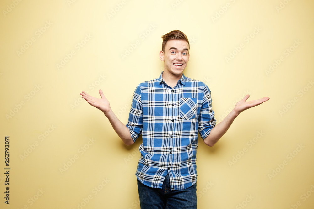 Cheerful guy dressed in a plaid shirt and jeans stands next to yellow wall in the studio Cheerful guy dressed in a plaid shirt and jeans stands next to yellow wall in the studio