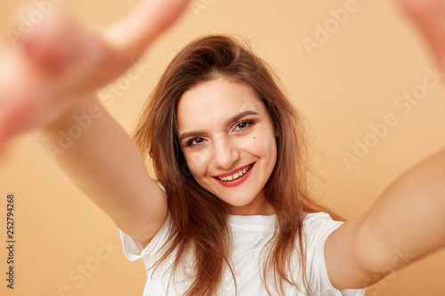 Brunette girl with long hair dressed in white t-shirt makes a selfie on the beige background in the studio