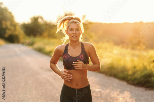 Beautiful dedicated blonde woman with ponytail and in sportswear running in nature on sunny summer day. Waist up.