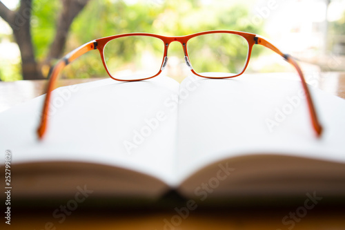 Orange eyeglasses with white notebook on wooden table, Bokeh garden background, Close up & Macro shot, Selective focus, Stationery concept