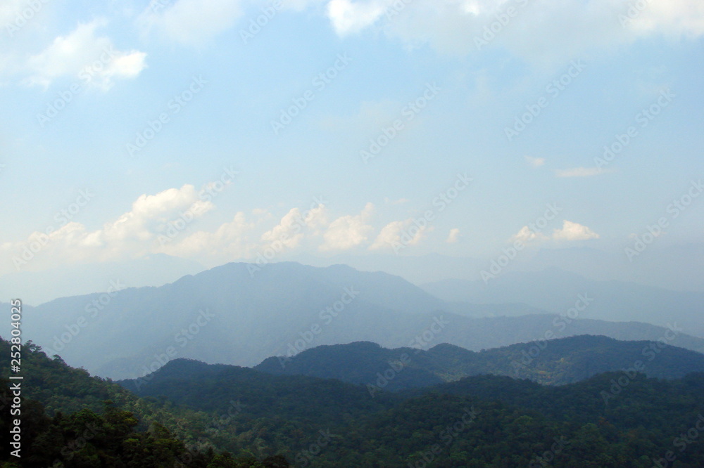 Panorama from a bird's eye view of a cloudy blue sky on the background of numerous Asian mountain ranges on a horizon covered with dense forests.