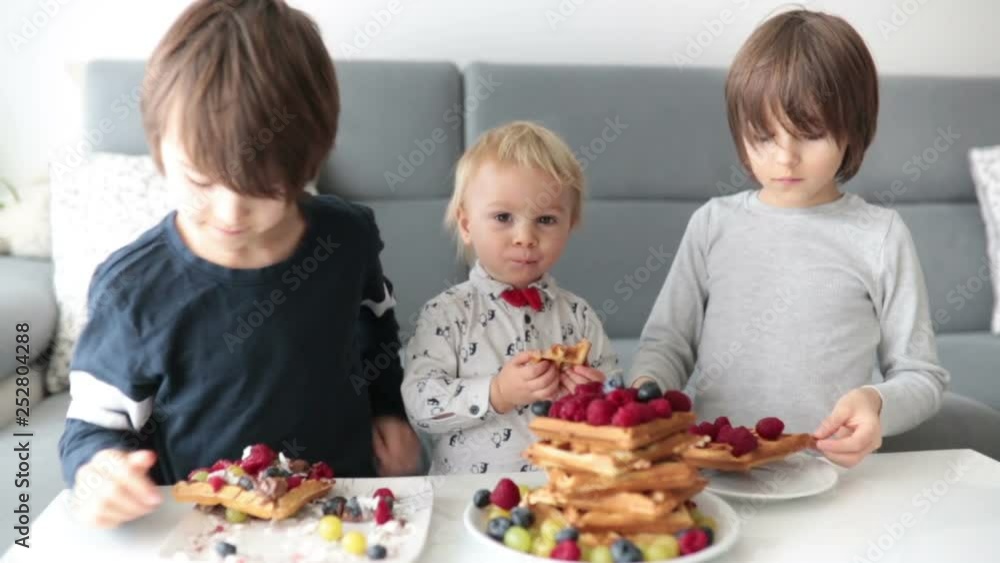 Sweet toddler birthday boy and his older brothers, eating belgian waffle with raspberries, blueberries, cocnut and chocolate at home Sweet toddler birthday boy and his older brothers, eating belgian waffle with raspberries, blueberries, cocnut and chocolate at home