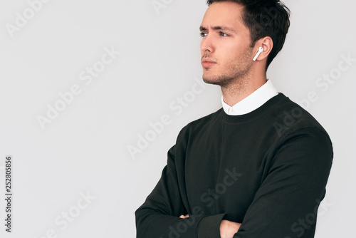 Serious young man standing with crossed hands and having a call via wireless earphones with a colleague isolated on white wall. Businessman using wireless earbuds for communication. Technology, people