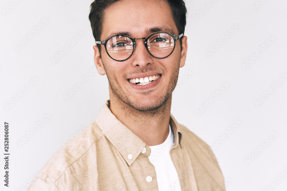 Close up portrait of cheerful young man, looks directly at the camera, wears round spectacles, isolated over white wall. Smart bristle student male smiling and wearing casual outfit. People emotions