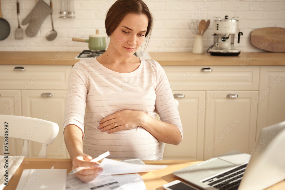 Indoor shot of busy serious young pregnant woman keeping one hand on belly, holding sheet of paper, paying bills online using laptop computer, having focused concentrated look, sitting in kitchen Indoor shot of busy serious young pregnant woman keeping one hand on belly, holding sheet of paper, paying bills online using laptop computer, having focused concentrated look, sitting in kitchen
