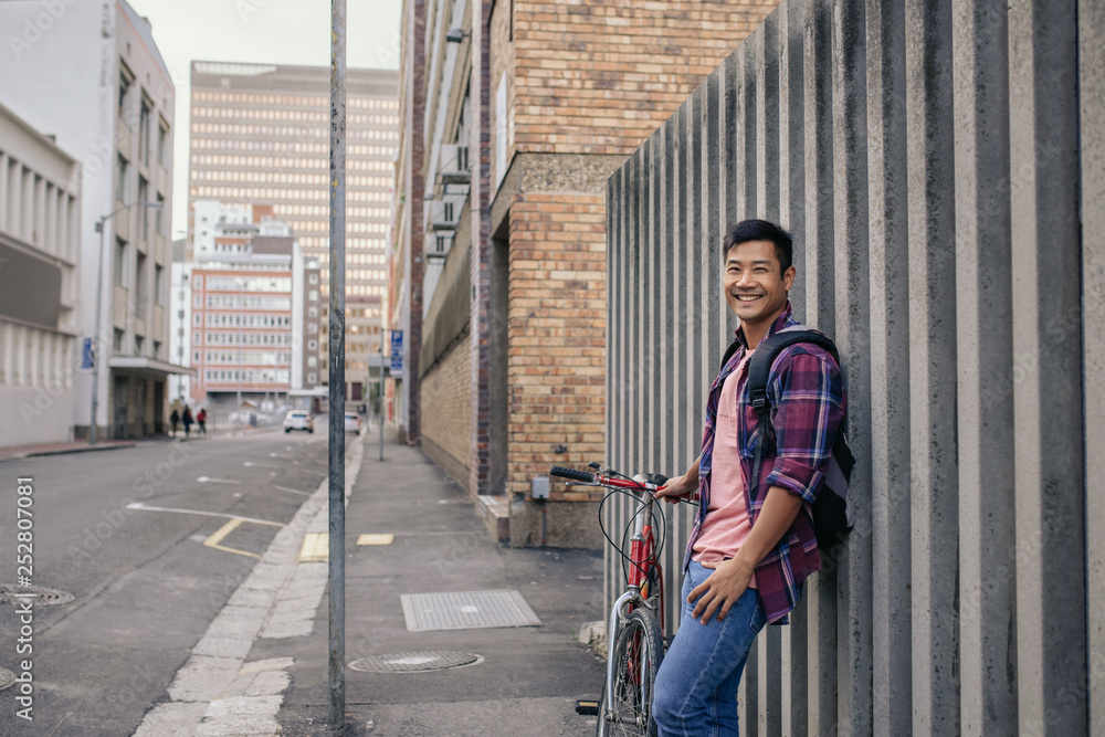 Smiling young man leaning with his bike against a wall