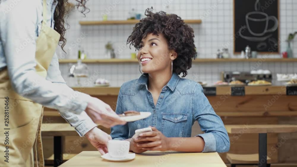 female waitress bringing coffee and cookies to beautiful smiling african american female customer in cafe female waitress bringing coffee and cookies to beautiful smiling african american female customer in cafe