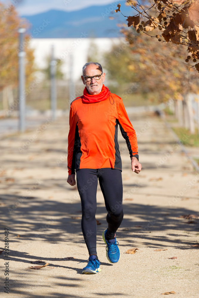 Front view of a senior man in sport clothes jogging in a city park in a sunny day