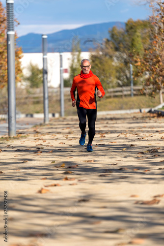 Front view of a senior man in sport clothes jogging in a city park in a sunny day