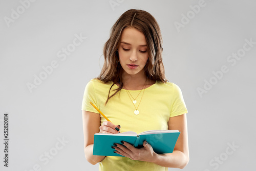 education, school, inspiration and people concept - young woman or teenage student girl in yellow t-shirt writing to diary or notebook by pencil over grey background