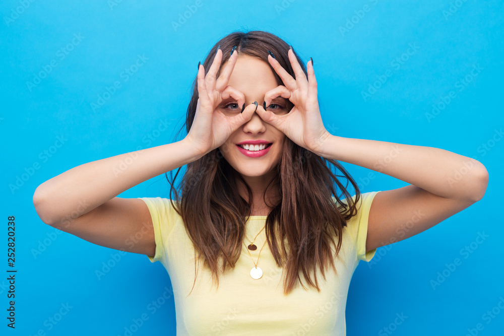 fun and people concept - smiling young woman or teenage girl looking through finger glasses over bright blue background fun and people concept - smiling young woman or teenage girl looking through finger glasses over bright blue background
