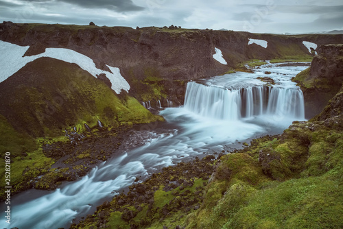 The Axlarfoss Waterfall with golden clouds in the sky. The flowing water is captured by a long exposure. Amazing blue color of water from the glacier. Natural and colorful environment...
