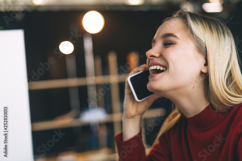 Cheerful young blonde woman in room laughing. He hold phone and talk. Horizontal portrait. Blurred background