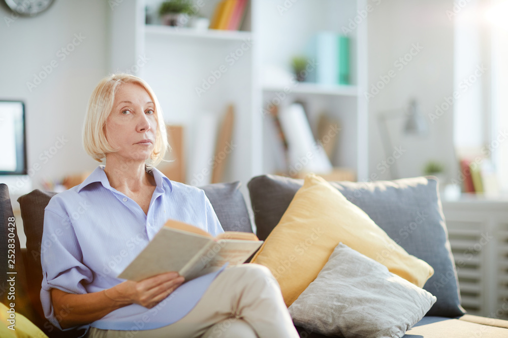 Portrait of contemporary adult woman reading book sitting on sofa at home and looking at camera, copy space Portrait of contemporary adult woman reading book sitting on sofa at home and looking at camera, copy space