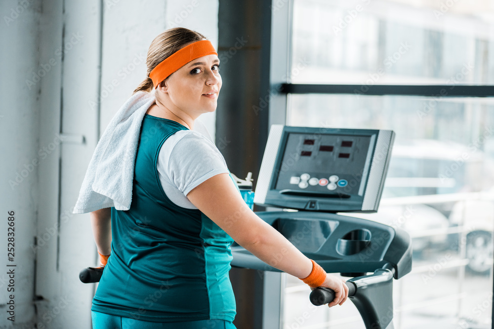 exhausted overweight girl with towel on shoulders looking at camera on treadmill in gym