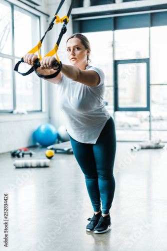 concentrated overweight girl training arms with suspension straps in gym photo