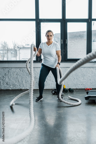 concentrated overweight woman training with battle ropes in gym