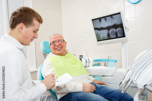 Happy and smile senior man 70-75 years old with white teeth on review of a dentist, sitting in chair dental clinic. Looking on young dentist doctor. Dental care for older people. Health care concept