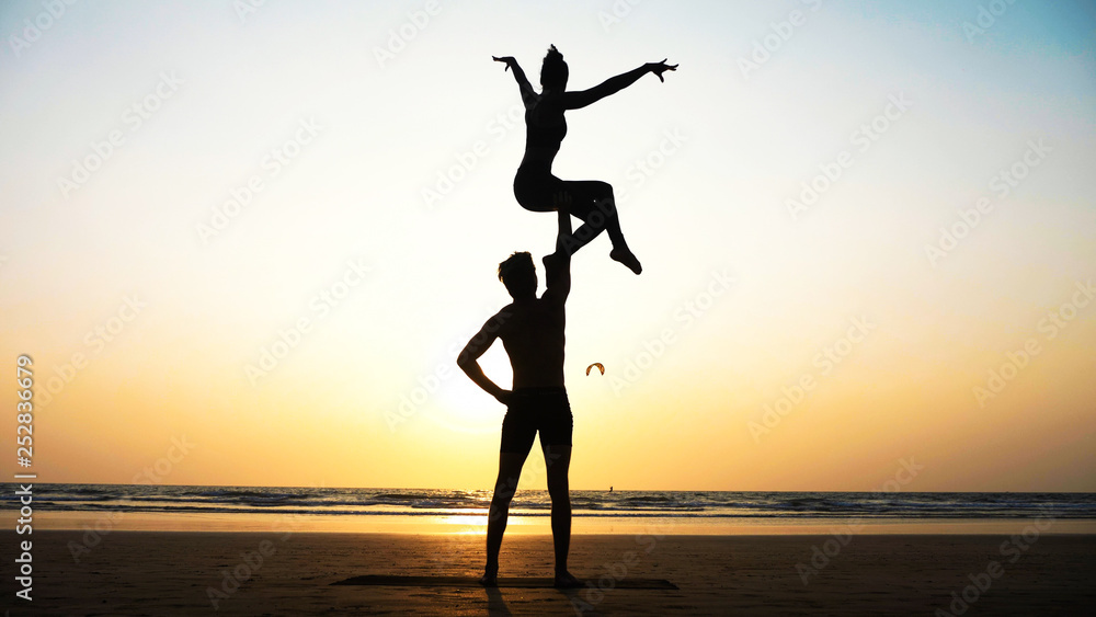 Silhouette of fit sporty couple practicing acrobatic yoga with partner together on the beach. Male standing and holding his female partner over his head. Mix race couple doing acrobatic exercise. 