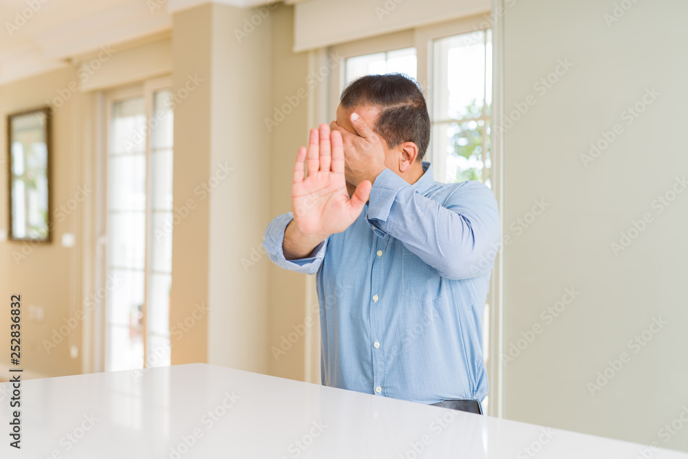 Middle age man sitting at home covering eyes with hands and doing stop gesture with sad and fear expression. Embarrassed and negative concept.