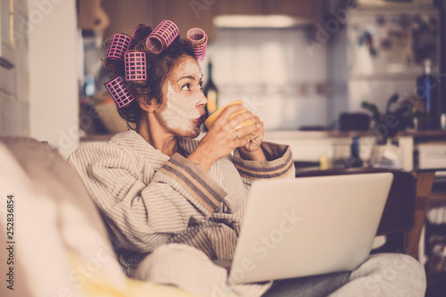 Funny housewife at home with pink curlers drinking a cup of tea while use a laptop on the sofa - technology and daily lifestyle at home - vintage filter colors and middle age lady enjoying photo