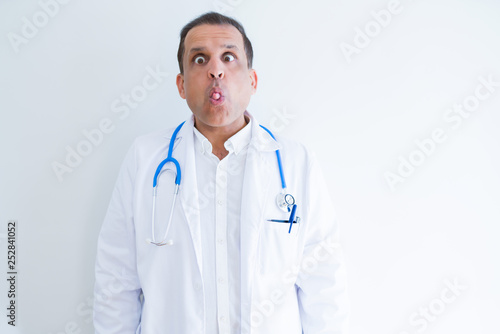 Middle age doctor man wearing stethoscope and medical coat over white background making fish face with lips, crazy and comical gesture. Funny expression.